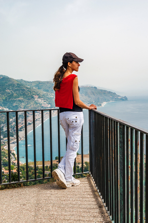 Girl enjoying the scenic view at Taormina, Sicily, standing on a platform with a rail and the mediterranean sea belowの写真素材