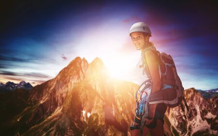 Fit athletic young woman out mountaineering standing in her gear backlit by the golden glow of the sun rising over a steep rocky mountain peak in a colorful sky turning to smile at the cameraの写真素材