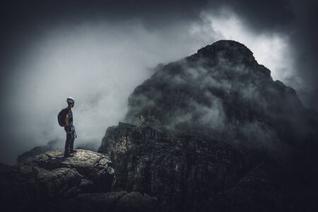 A mountain climber standing on a peak with a view over a dark, ominous, stormy mountain range.の写真素材