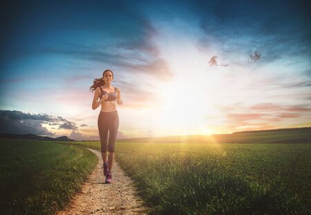 Healthy fit young woman jogging at sunrise running along a footpath through green fields backlit by...