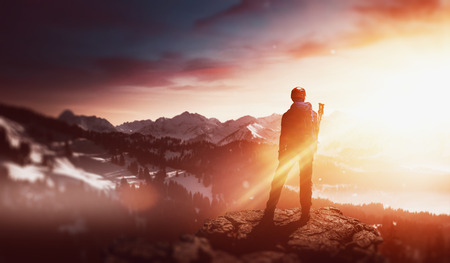 Hiker standing on a rocky mountain summit watching the golden glow of the rising sun at dawn over a winter landscape of snowy mountain peaks and valleys in an atmospheric panoramaの写真素材