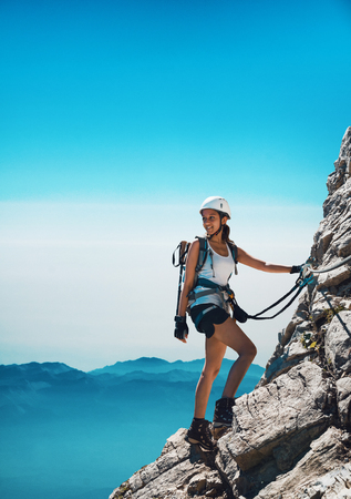 Fit attractive woman mountaineer attached to ropes standing on a rocky outcrop on a cliff looking back at the valley behind her with a happy smile of pleasureの写真素材