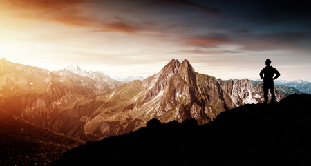Man standing silhouetted on a rocky ledge watching the sun rise over rugged mountain peaks and...