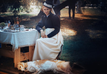 The photo shows a woman in a medieval costume sitting beside a set table outdoors, with a dog lying nearby. Atmosphere of historical reenactment.の写真素材