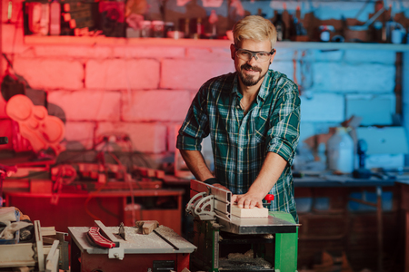 Attractive young hipster bearded man with eye protectors by profession carpenter builder saws with a circular saw a wooden board on a wooden table in the workshop.の写真素材