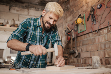 Attractive blonde young hipster bearded man by profession carpenter builder nailing wood board with...