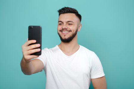 Handsome young bearded hipster in white t-shirt using phone and taking selfie with bright smile.の写真素材