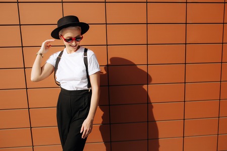 Attractive trendy hipster young woman with stylish red sunglasses and black hat stay near wall.