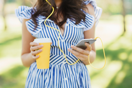 Close-up photo of young woman in summer clothes listening to music with headphones while holding mobile phone and cup of coffee in the park.の写真素材