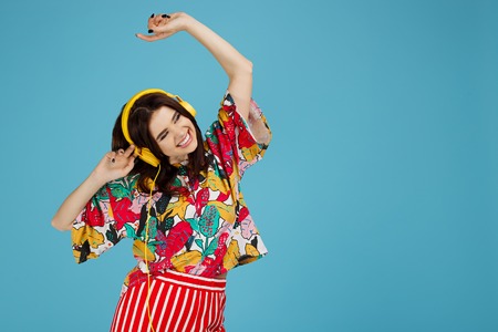 Photo of happy brunette woman in colorful casual outfit with yellow headphones listening music on the blue background. Bright young woman danceの写真素材