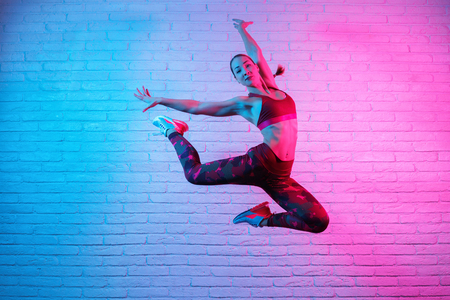 Charming young slim gymnast woman in sports clothing exercises in against brick wall in neon lights. Flexible muscular woman doing gymnastic elements.の写真素材
