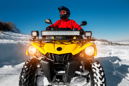 Photo of attractive young man in red warm winter clothes and black helmet on the ATV 4wd quad bike stand in heavy snow with deep wheel track. Moto winter sports.の写真素材