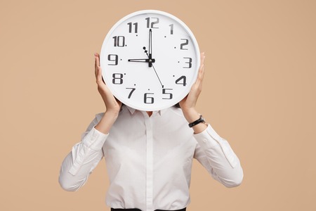 Photo of young modern business woman hold a clock in front of the face over beige background.の写真素材