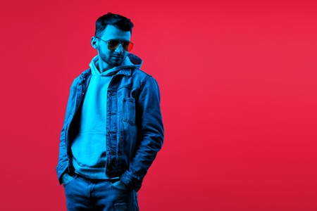 Cheerful stylish hipster bearded young man wearing yellow sunglasses and blue jeans jacket posing in...