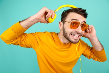 Cheerful bearded young man wearing yellow sunglasses listening to music with yellow headphones on cyan background.の写真素材