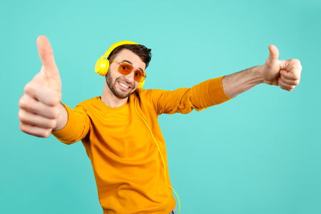 Awesome funny bearded young man wearing yellow sunglasses listening to music with yellow headphones showing ok gesture over cyan background.の写真素材