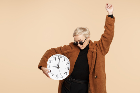 Attractive young hipster woman with blonde short hair wearing a coat and sunglasses holding a clock isolated over beige background.の写真素材