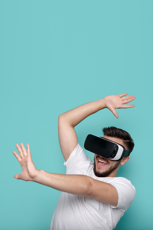 Shocked handsome bearded man wearing virtual reality goggles in studio over cyan background....