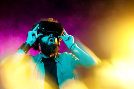 Young shocked bearded hipster man with curly hair wearing virtual reality goggles in studio in neon...