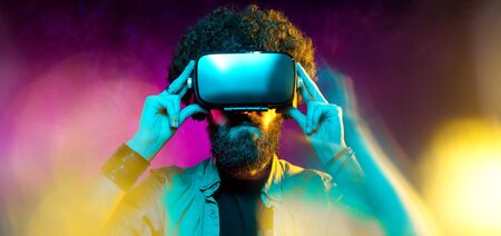 young bearded hipster man with curly hair wearing virtual reality goggles in studio in neon lights....