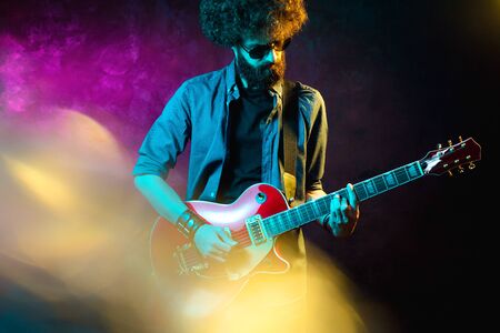 Young hipster man with curly hair with red guitar black sunglasses in neon lights. Rock musician is playing electrical guitar.の写真素材
