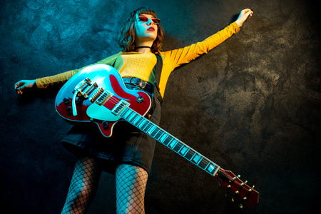Charming hipster woman with curly hair with red guitar in neon lights. Rock musician is playing electrical guitar. 90s style concept.の写真素材