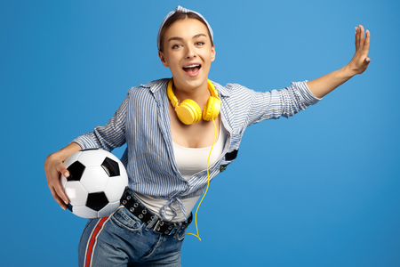 Photo of adorable young woman with yellow penny or skateboard, soccer ball and headphones dance over blue background.の写真素材