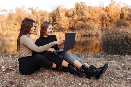 Lovely two young twin sisters in casual outfit with bright smile pointing on notebook or laptop screen at the parkの写真素材