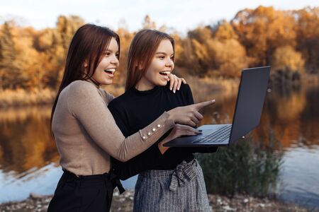 Attractive two young twin sisters in casual outfit with bright smile pointing on notebook or laptop screen at the parkの写真素材