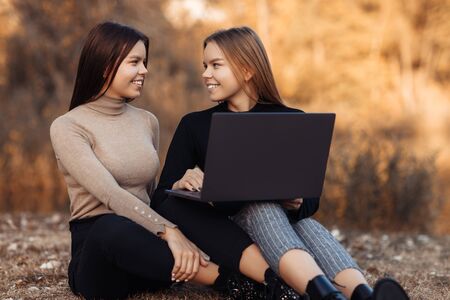 Funny two young twin sisters in casual outfit with bright smile use notebook or laptop at the park