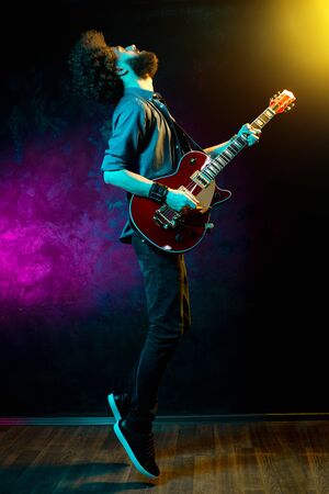 Young hipster man with curly hair jump with red guitar in neon lights. Rock musician is playing electrical guitar.の写真素材