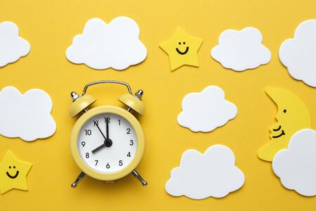 Yellow round alarm clock, moon, stars and white clouds on the yellow background.
