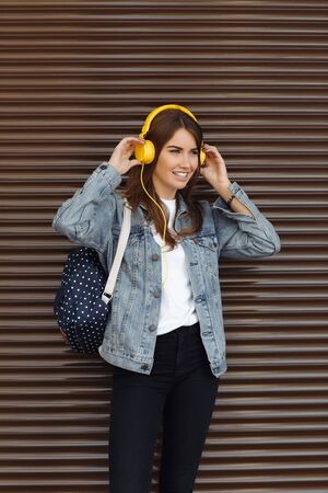Pretty smiling girl with brown hair wearing a backpach and yellow heaphones outdoors. Young woman listening music.の写真素材
