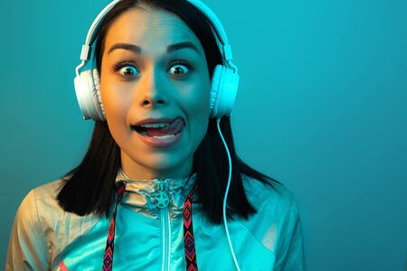 Shocked young woman in 90s style sport outfit with white headphones listening to music in neon...