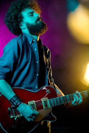 Hipster man with curly hair play with passion red guitar in neon lights. Rock musician is playing electrical guitar.の写真素材