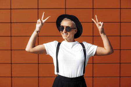 Pretty hipster young woman wearing red sunglasses and black hat near wall showing peace gesture hand sign.の写真素材