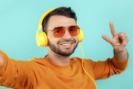 Trendy bearded young man wearing yellow sunglasses listening to music and take sefie over cyan...