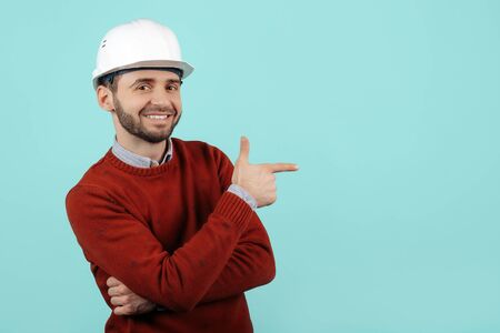 Handsome young bearded engineer or constructor man in casual outfit point finger away over cyan...
