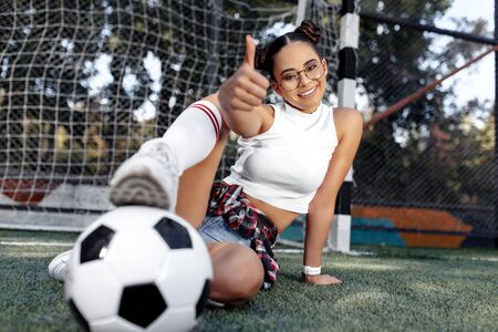 Funky young teenage hipster girl sitting on the grass showing thumb up in front of goal post at the stadium.の写真素材
