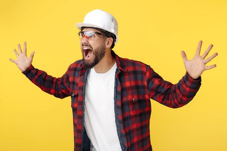 Excited bearded engineer or constructor man in plaid shirt holding hands up and screaming over yellow background.の写真素材