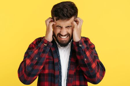 Bearded hipster young man in plaid shirt suffer headache rubbing temples keeping eyes closed over...