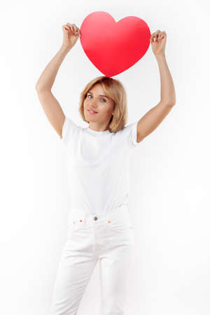 Charming young blonde woman in casual white outfit holding paper heart on the head over white background.の写真素材