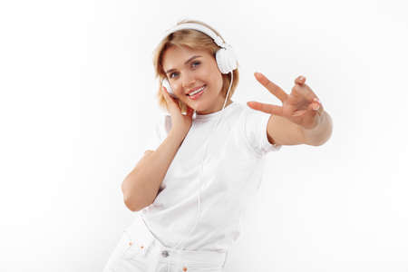 Positive young blonde woman in casual white outfit listening music in headphone over red background....