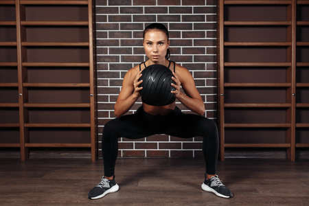 Front image of fit young brunette woman doing squat exercise with medicine ball in gym.