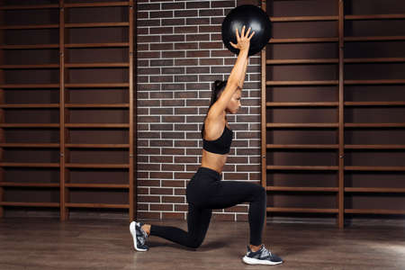 Fit young brunette woman doing squat exercise with medicine ball in gym.