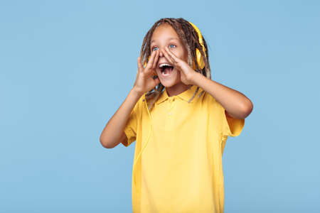 Funny little boy with african dreads listening music in headphones screaming over blue background.の写真素材