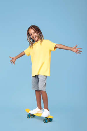 Gleeful little boy with african dreads on skateboard having fun over blue background.