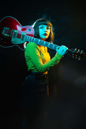 Hipster woman with curly hair with red guitar in neon lights. Rock musician with electrical guitar. 90s style concept.の写真素材