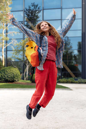 Full lengh image of stylish young blonde hipster woman student with backpack jumping outdoors at...
