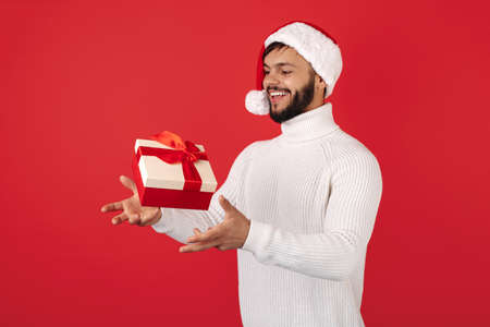 Funny hipster man wears santa claus hat cathing flying gift box over red background. Christmas and...